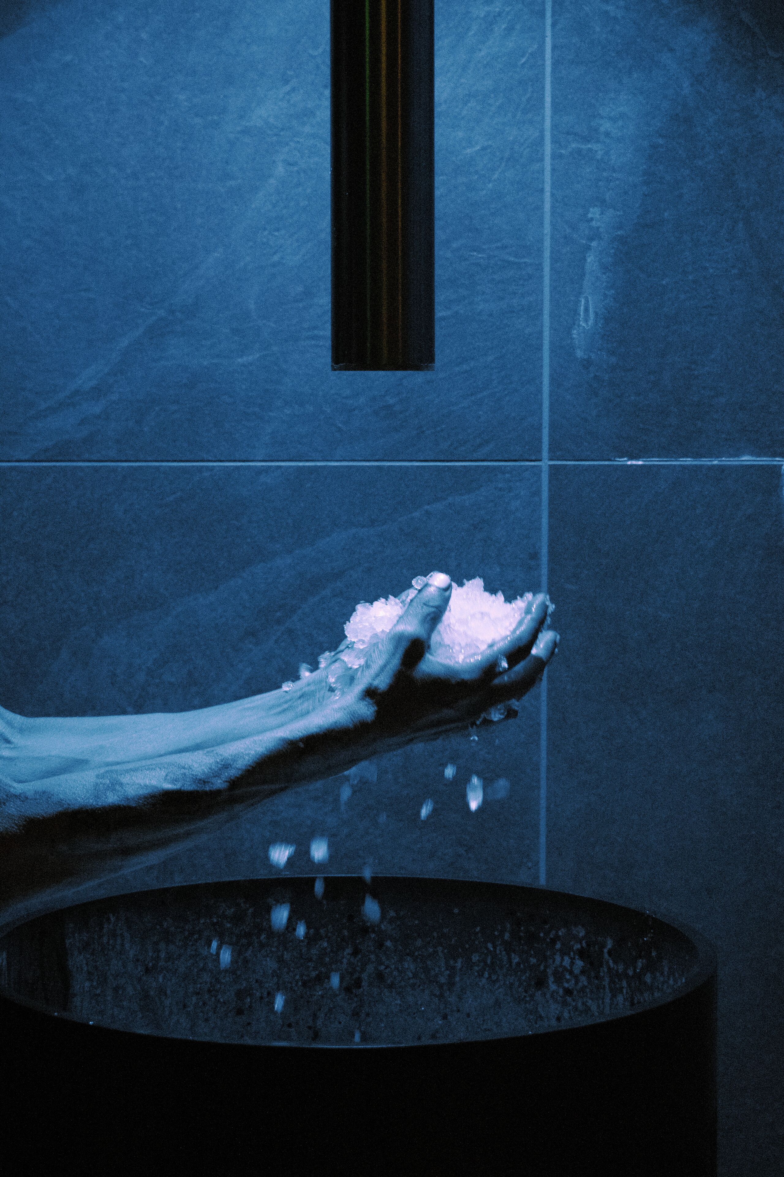 A hand holding a piece of ice over a dark, cylindrical container.