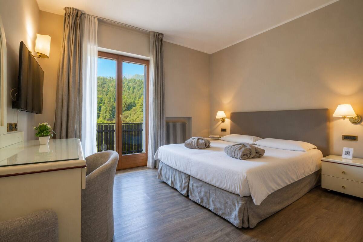 Una camera d'albergo con un letto, lampade sui comodini, una scrivania con lampada, una sedia e vista sul verde attraverso una finestra, oltre a un balcone.