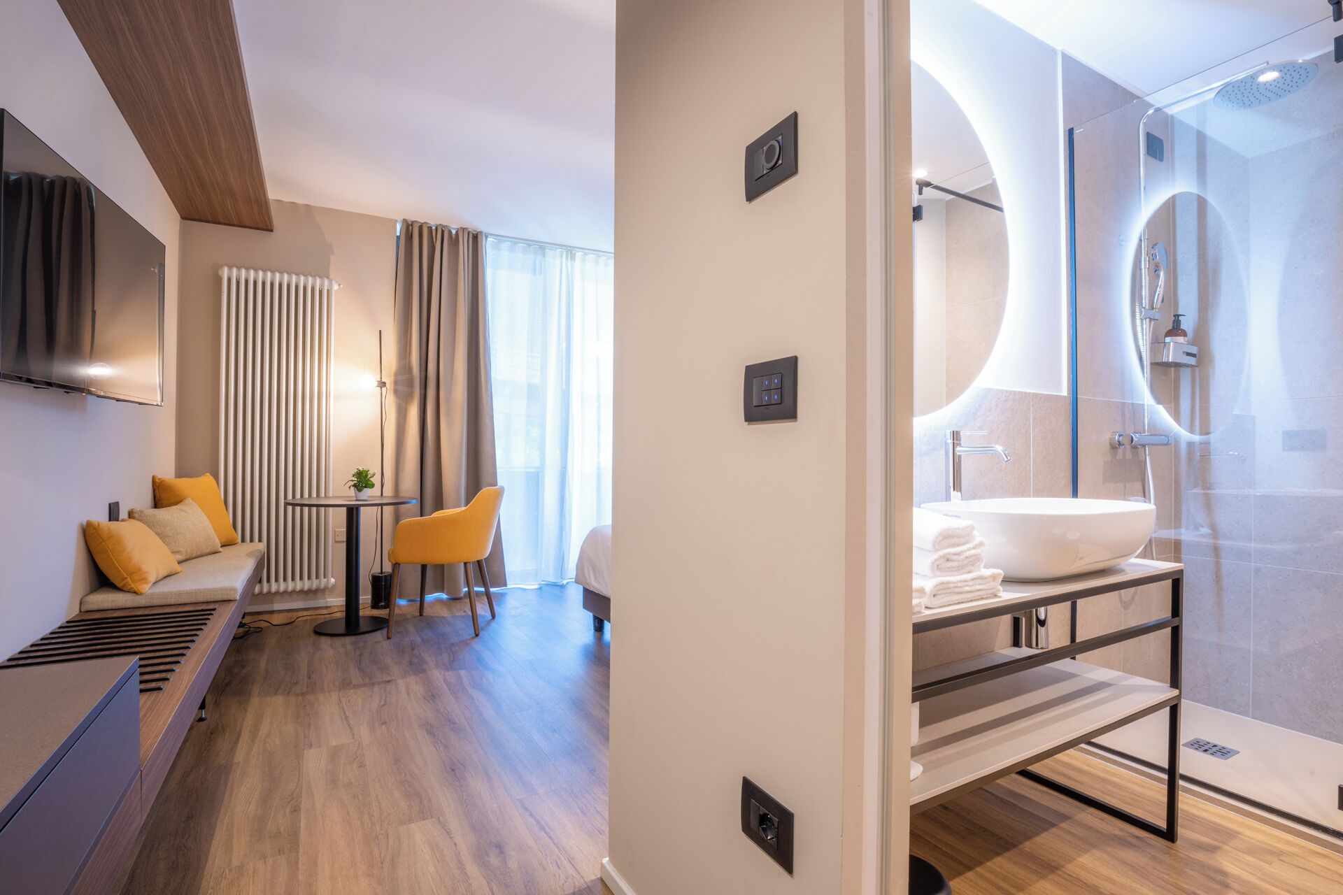 A modern hotel room with a sofa, a small table, a TV, a bathroom with a large sink and shower.