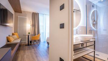 A modern hotel room with a sofa, a small table, a TV, a bathroom with a large sink and shower.