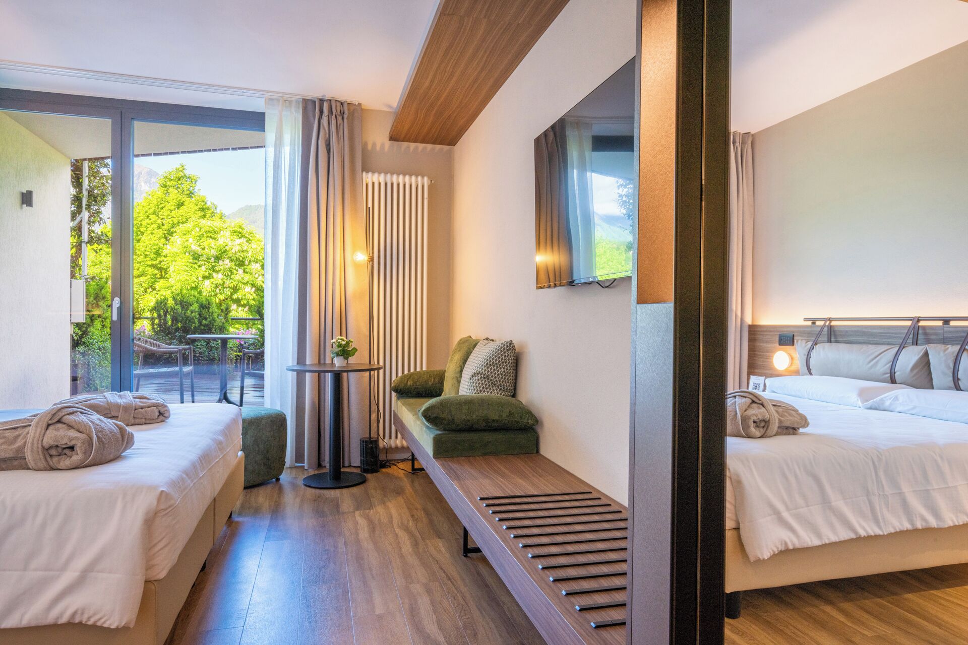 Una habitación de hotel moderna con dos camas, un banco, una mesita y vistas al exterior con vegetación.