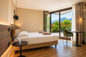 A modern hotel room with a bed, a small table, a chair, and a view of mountains through large windows. - Alchimia WellResort (Ponte Arche)