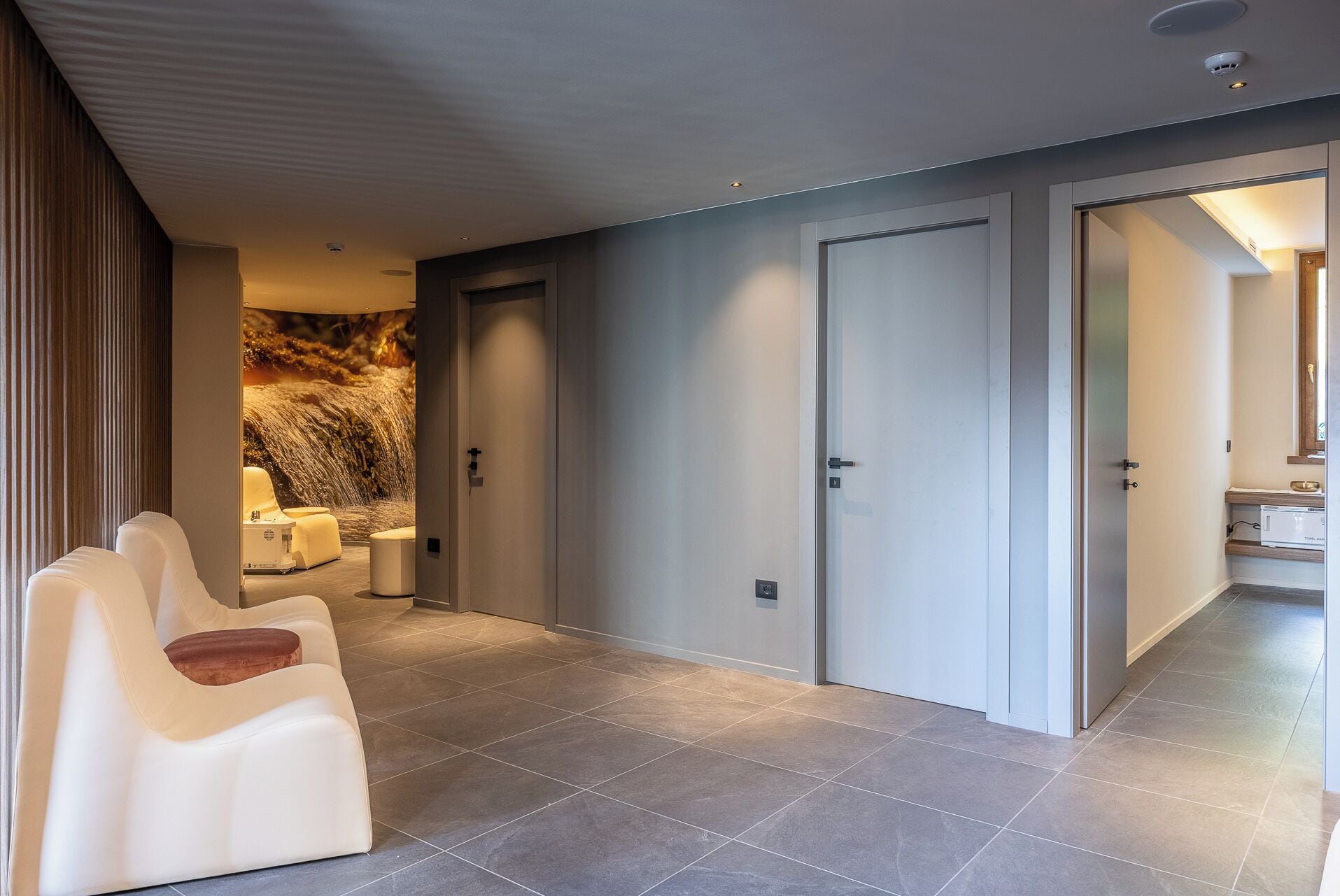 A modern hotel lobby with a seating area, a stone wall with a waterfall image, and a view into a bathroom.