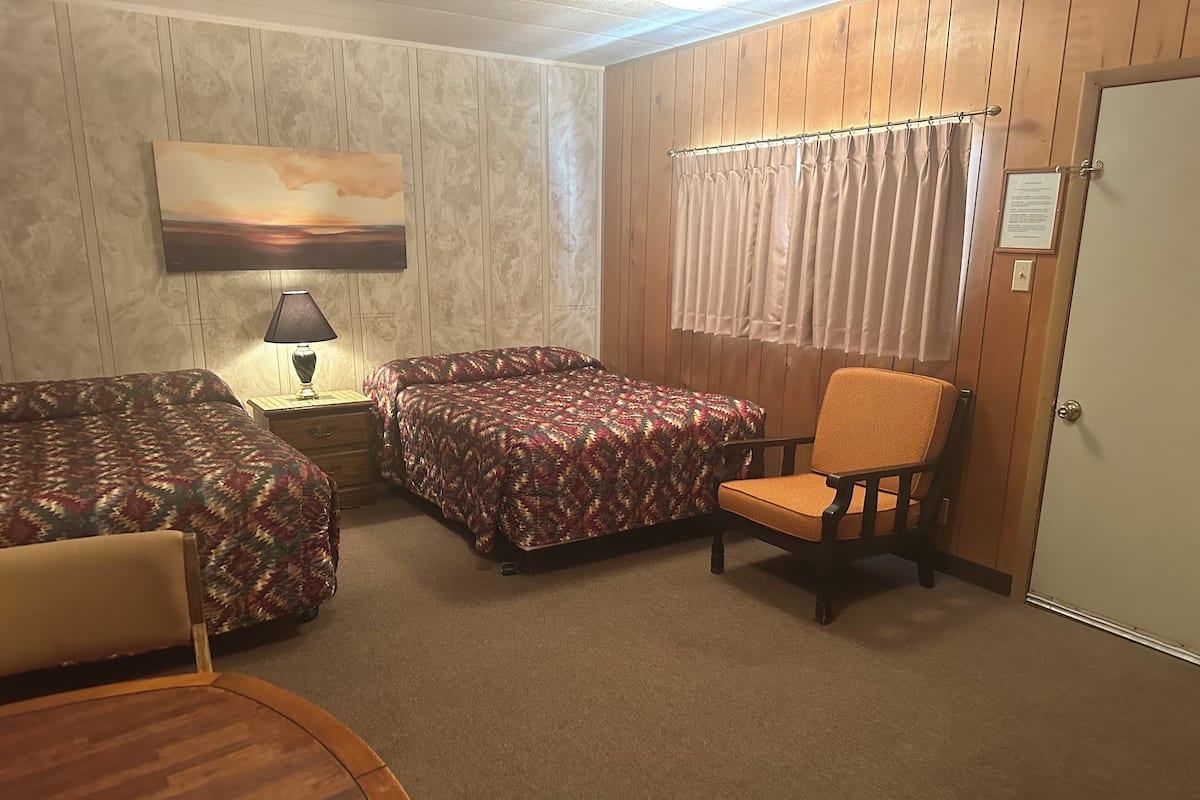 A room with two beds, a chair, a table, a lamp, and a painting on the wall.