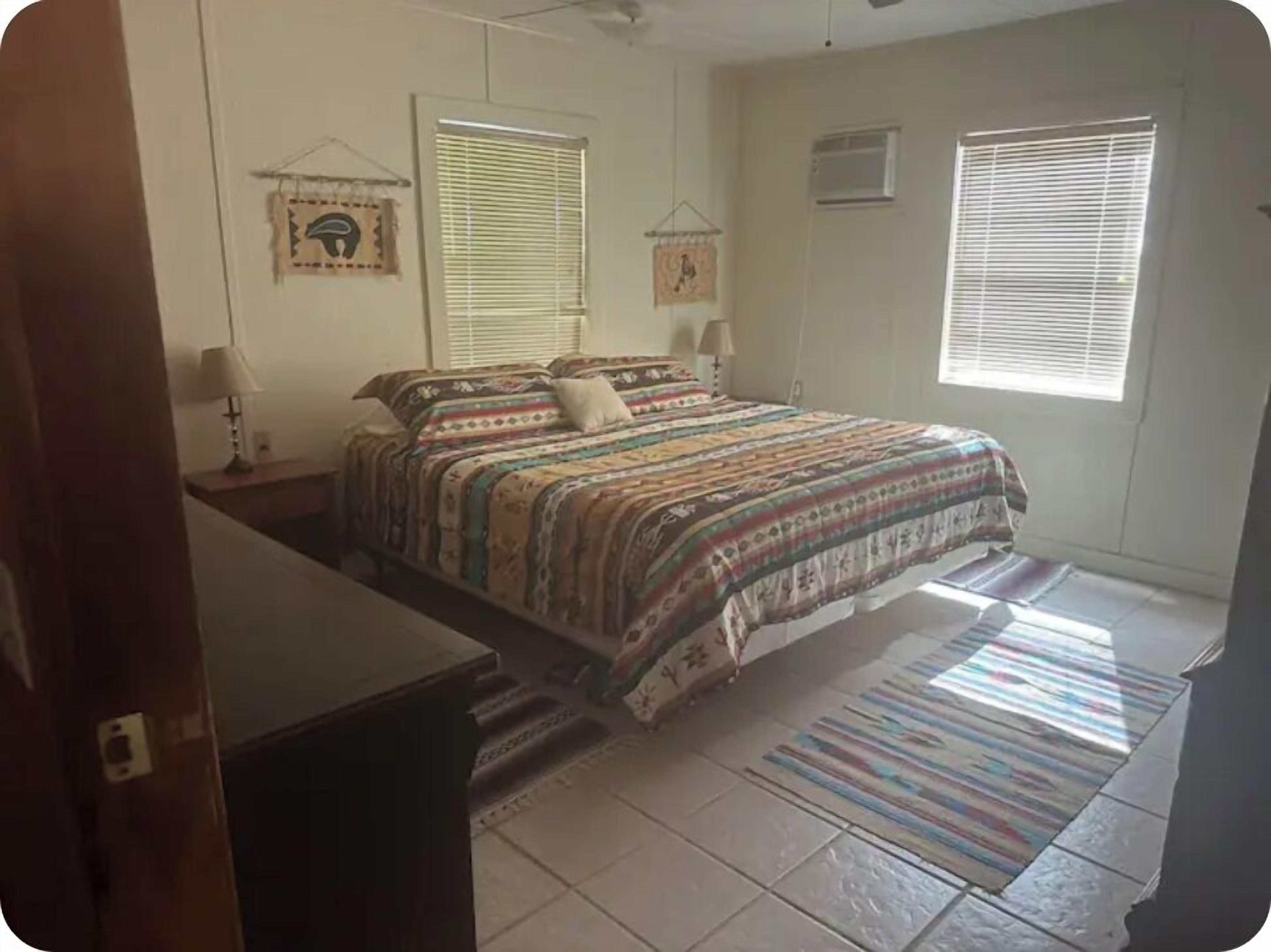 Un dormitorio con cama, dos mesitas de noche, una ventana con persianas y un aire acondicionado de pared, además de una alfombra estampada.