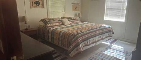 Un dormitorio con cama, dos mesitas de noche, una ventana con persianas y un aire acondicionado de pared, además de una alfombra estampada.