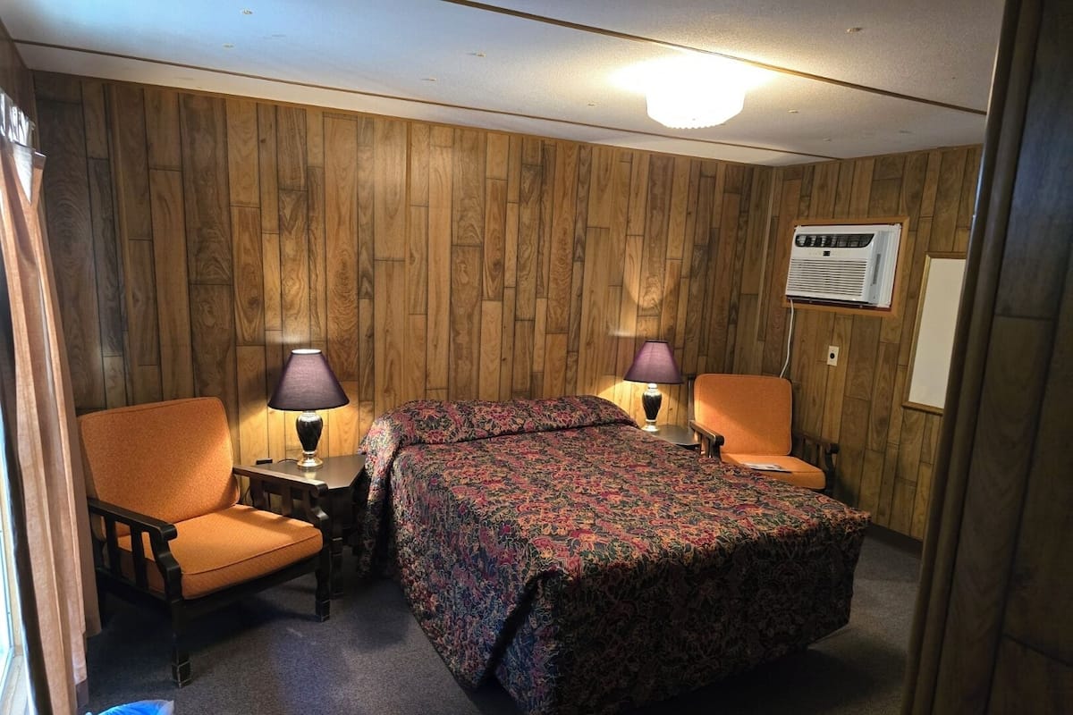 A room with a bed, two chairs, a lamp, and an air conditioning unit.