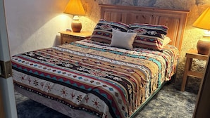 A bedroom with a stone wall, a wooden bed frame, a patterned bedspread, two bedside lamps, and a small wooden nightstand. - Chisos Mining Co Motel (Terlingua)