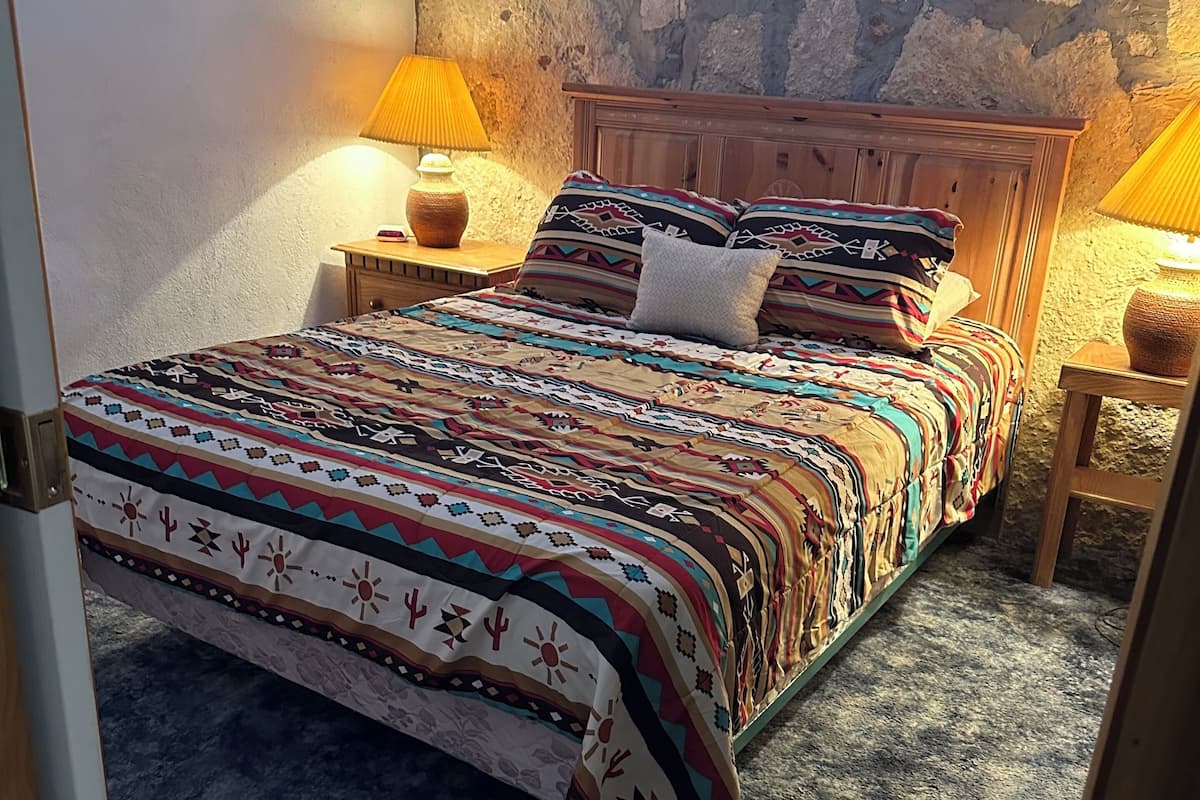 A bedroom with a stone wall, a wooden bed frame, a patterned bedspread, two bedside lamps, and a small wooden nightstand.
