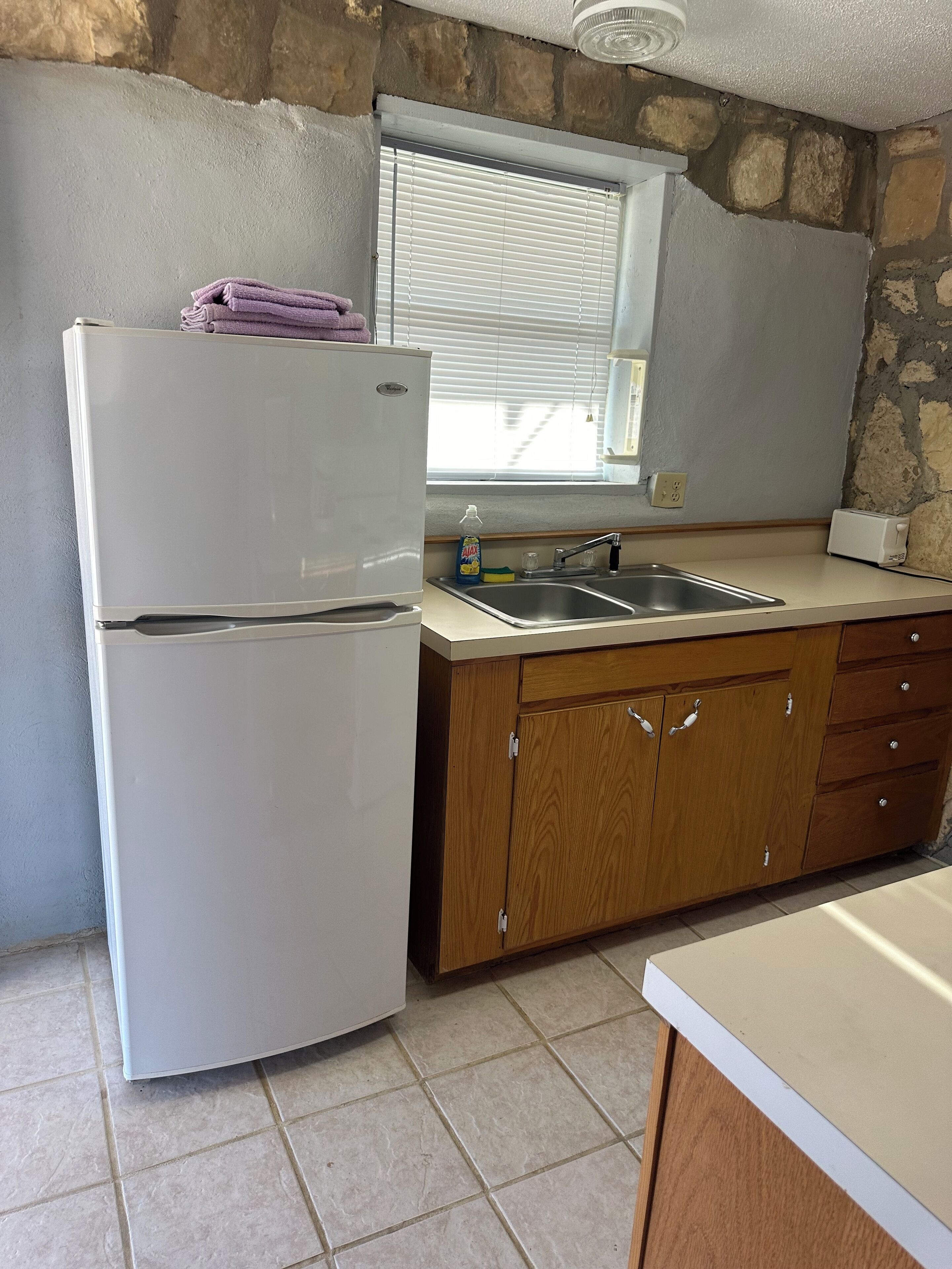 A kitchen with a white refrigerator, wooden cabinets, a window with blinds, and a stone wall.
