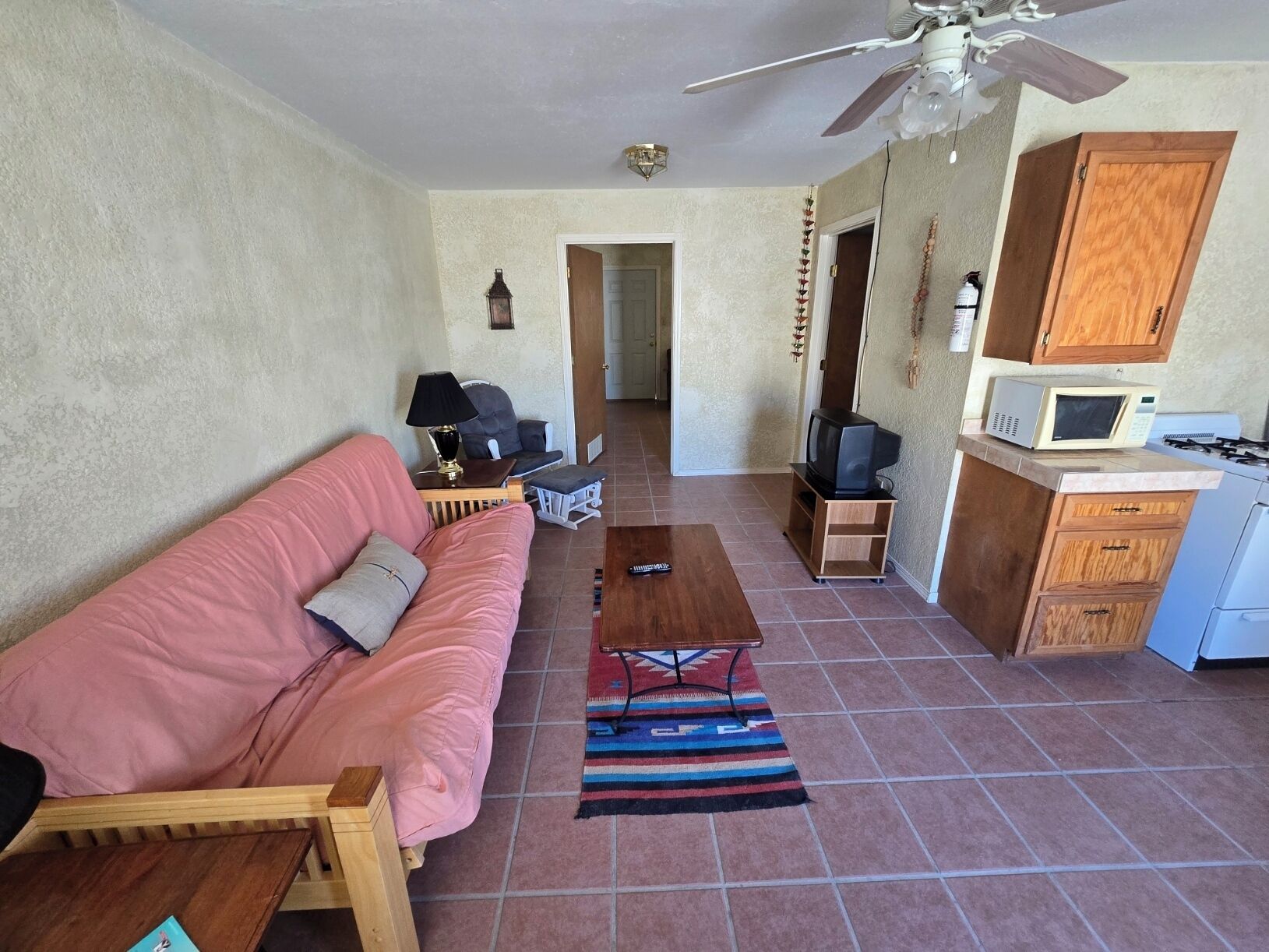Una sala de estar con un sofá rosa, una mesa de centro de madera, un televisor sobre un mueble de madera, un microondas en un armario y un ventilador de techo.