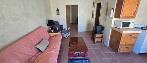 Una sala de estar con un sofá rosa, una mesa de centro de madera, un televisor sobre un mueble de madera, un microondas en un armario y un ventilador de techo.