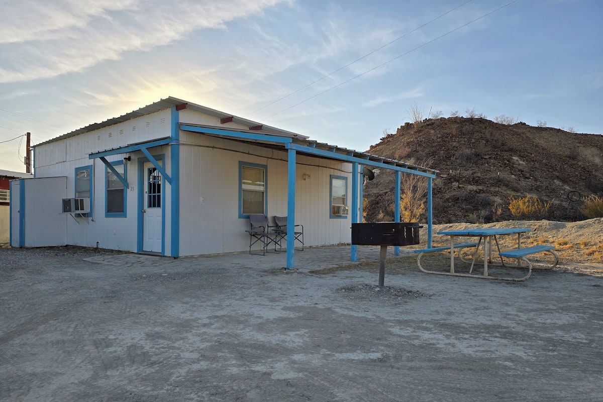 A small, single-story building with a blue and white exterior, a covered outdoor seating area, and a dirt ground.