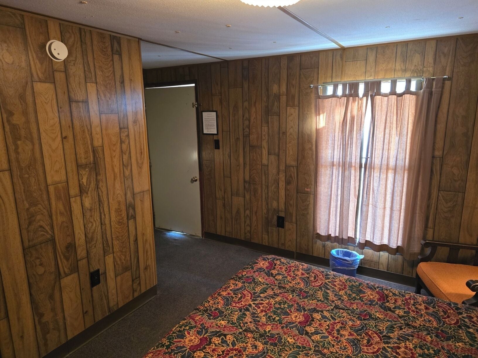 A room with wooden panel walls, a patterned bedspread, a door leading to another room, and a window with curtains.