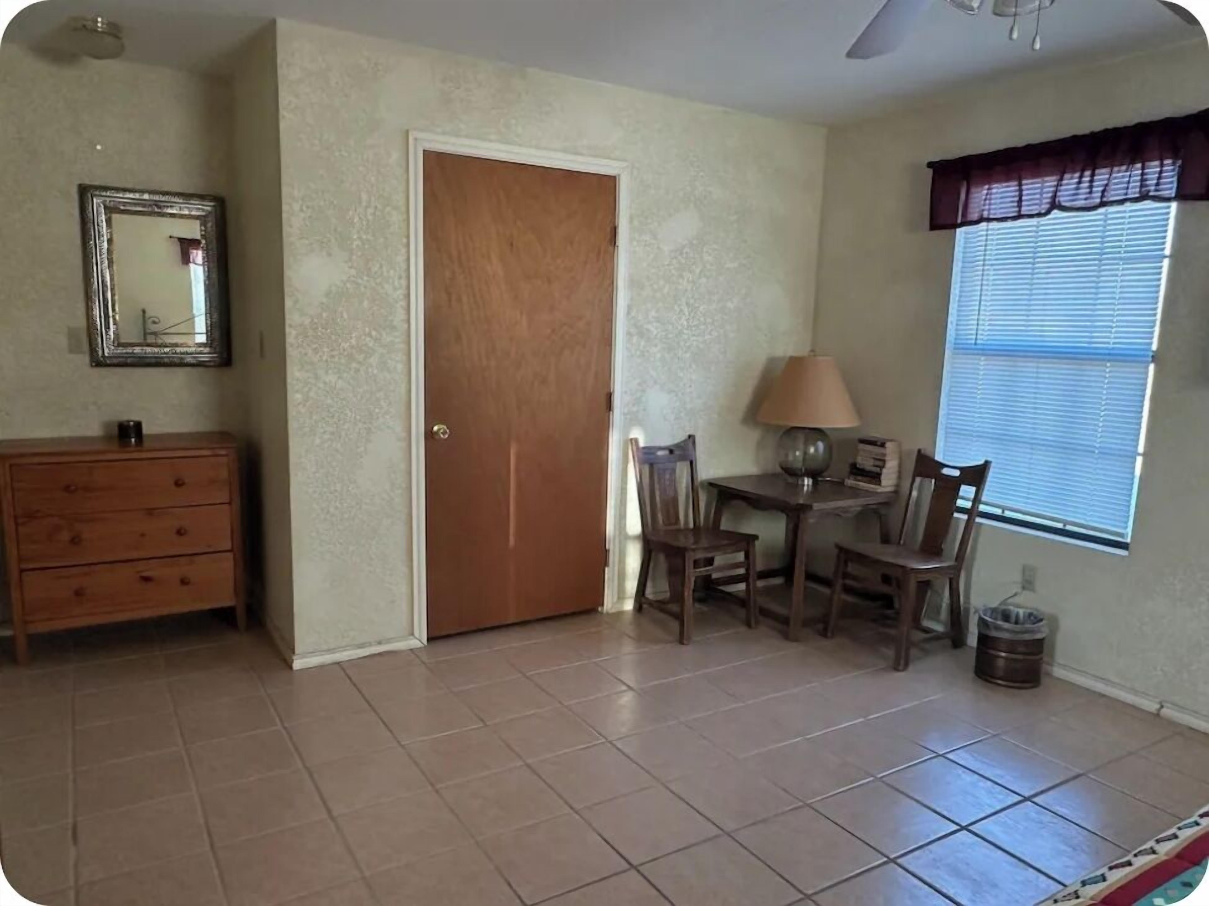 A room with a wooden dresser, a door, a table with chairs, a lamp, and a window with blinds.