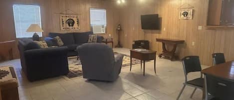 A living room with a sofa, armchairs, a coffee table, and a TV. There are wooden walls and a ceiling fan.