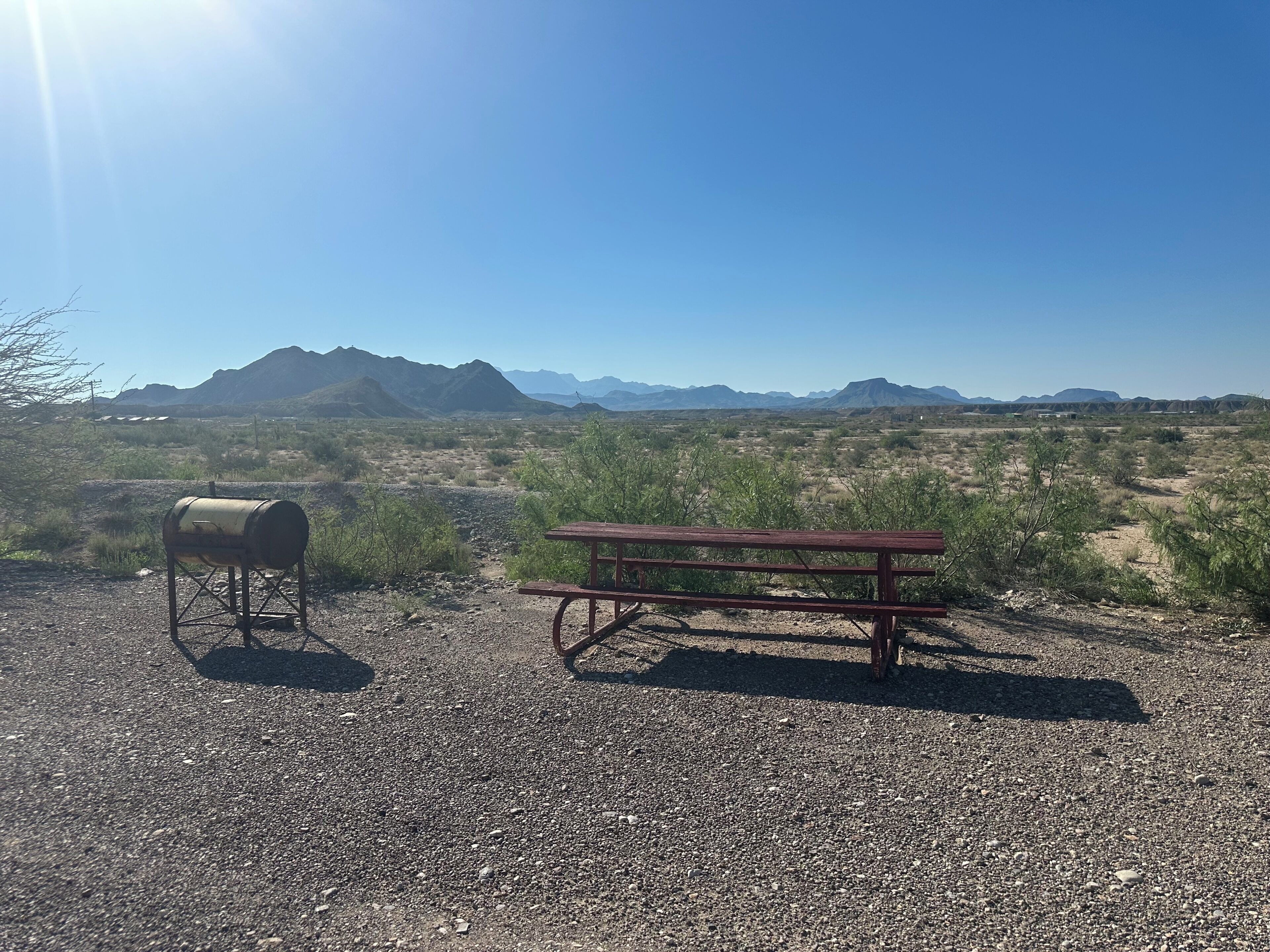 A gravel area with a wooden picnic table and a metal barrel, surrounded by sparse vegetation and distant mountains under a clear sky.