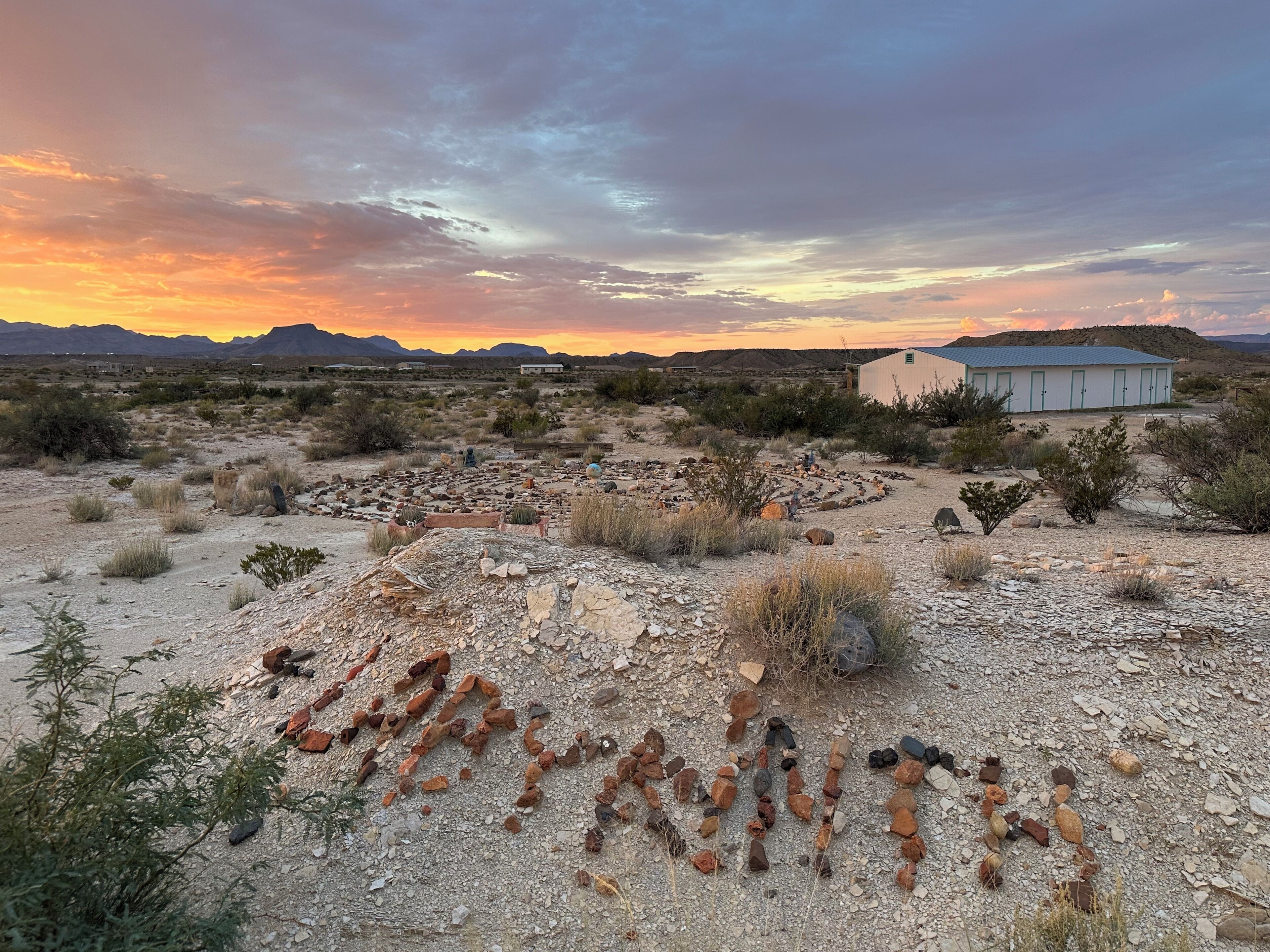 A desert landscape with scattered rocks and sparse vegetation, a small building in the distance, and a sunset sky.