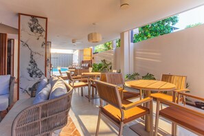 A modern outdoor dining area with wicker furniture and wooden tables, overlooking a pool and lush greenery. - Aquaraa (Maduvvari)