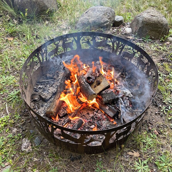 A metal fire pit with burning logs and embers, surrounded by grass and rocks.