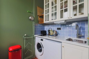 Cocina compacta con lavadora, lavamanos y un bote de basura rojo.