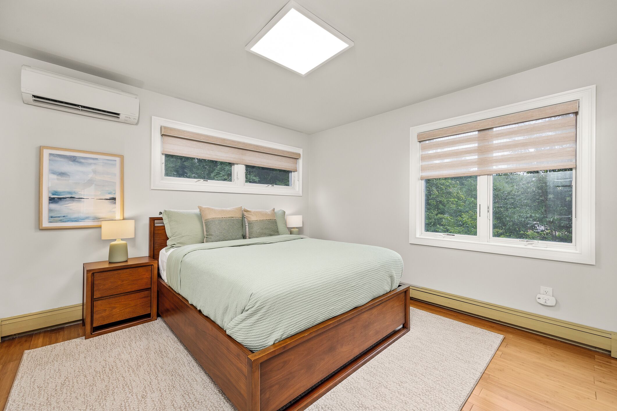 A bedroom with a bed, bedside tables, a nightstand, a window with blinds, and an air conditioning unit.