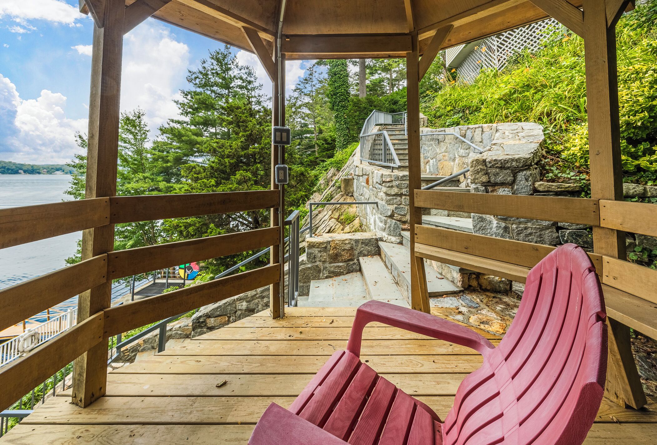 A wooden deck with a red Adirondack chair, stone steps, and a view of a lake and forest.