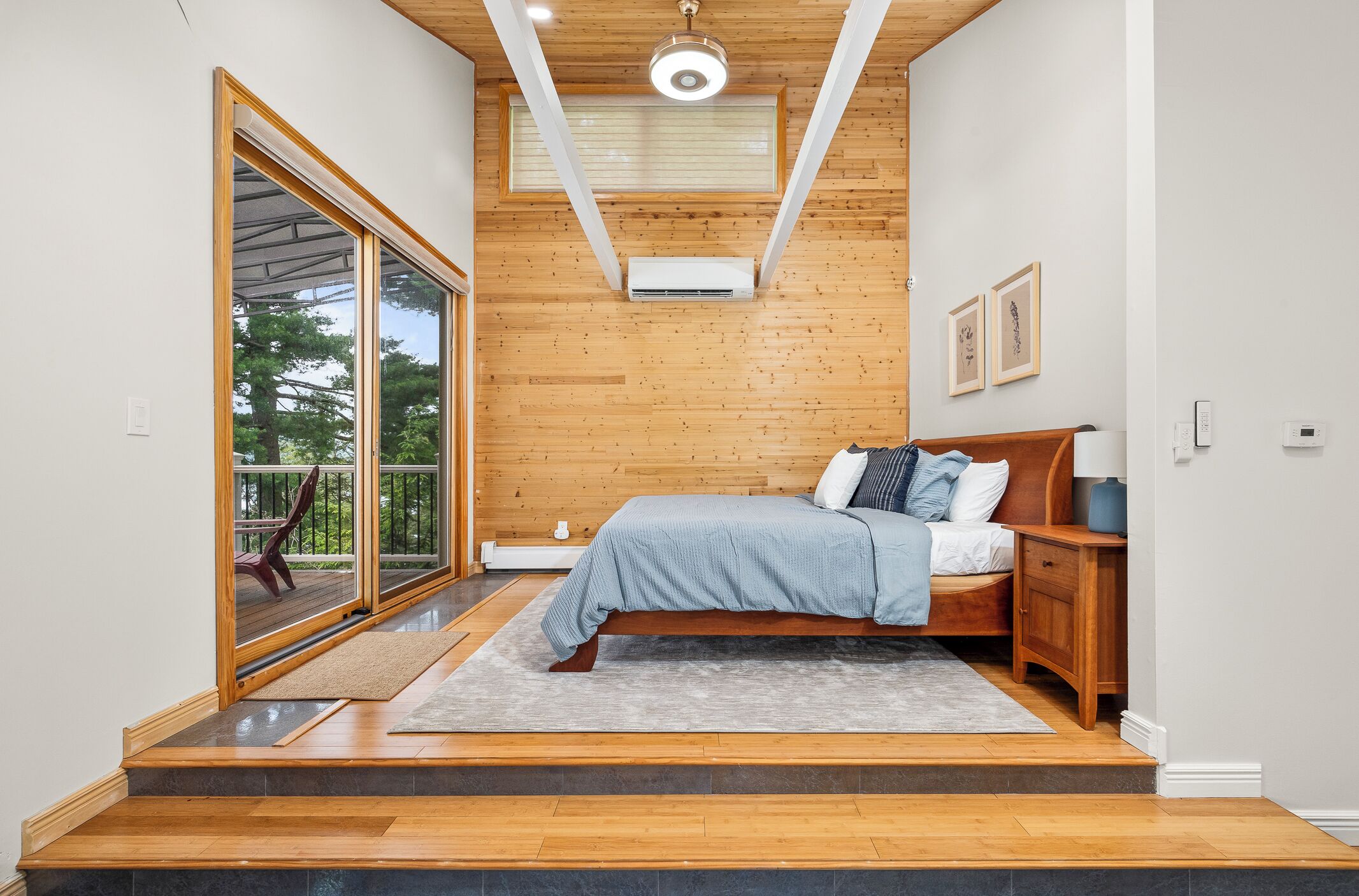 Une chambre avec une grande porte-fenêtre coulissante donnant sur un balcon, un plafond en bois et un lit recouvert d’une couverture grise.
