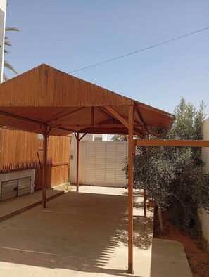 Un espacio exterior techado con techo de madera y vigas de metal, suelo de concreto y una puerta de garaje blanca al fondo.