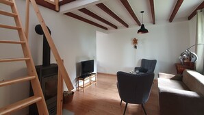 Ein Loft-Zimmer mit freiliegenden Holzbalken, einer Holzleiter, einem Fernseher, einem blauen Sessel und einem Sofa.