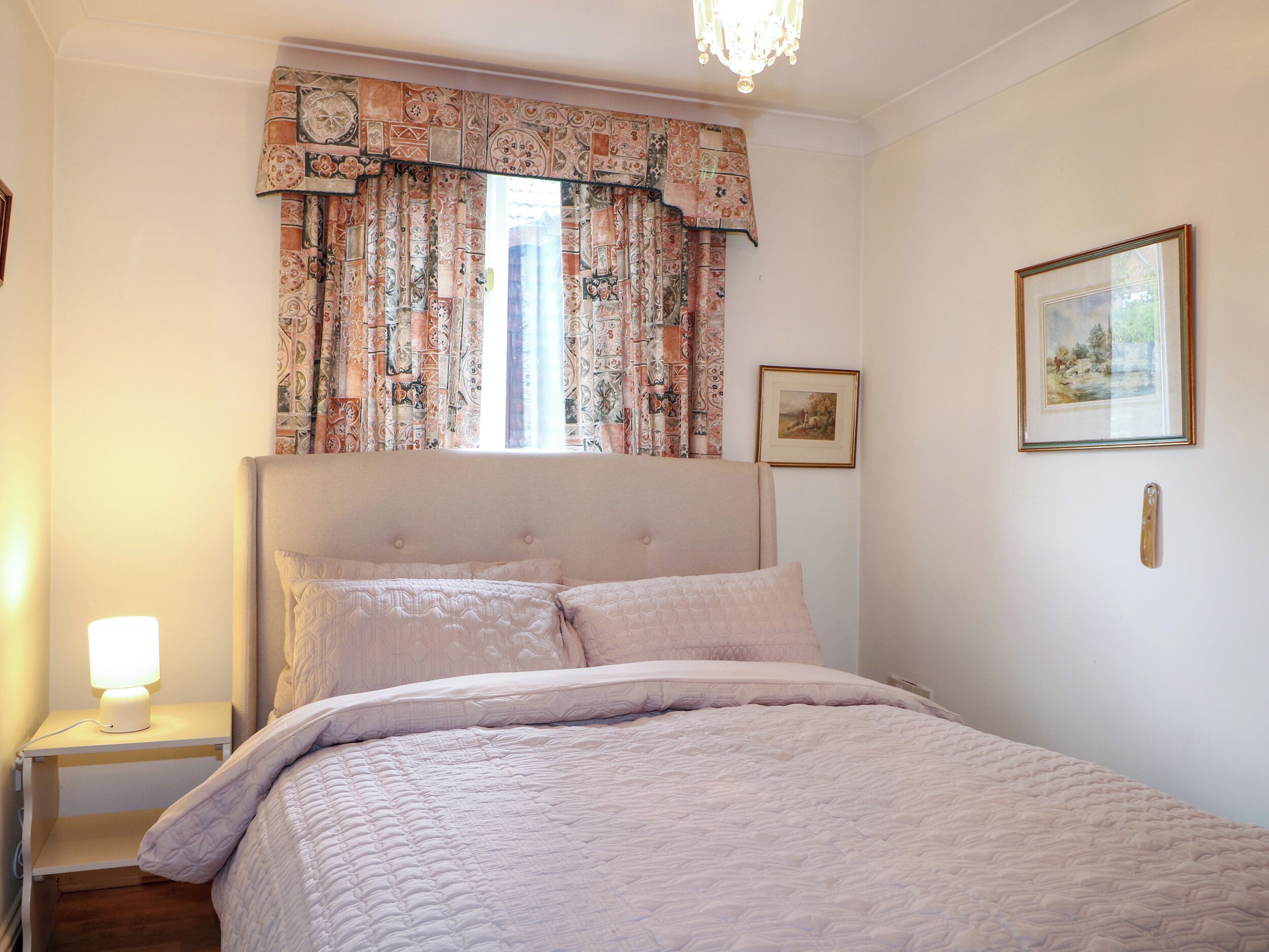 A bedroom with a bed, a nightstand, a lamp, a window with patterned curtains, and a framed picture on the wall.