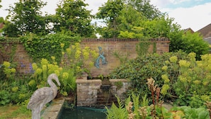 A garden with a stone swan statue, a brick wall with graffiti, and a variety of plants and flowers.