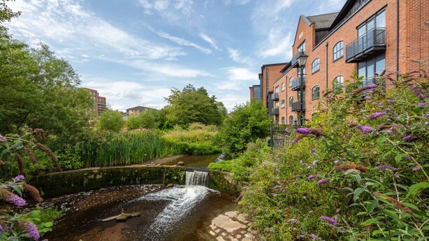 Un cadre paisible au bord de la rivière, agrémenté d’une petite cascade, d’une végétation luxuriante et d’un bâtiment en briques doté de balcons.