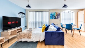 Une chambre d’hôtel moderne dotée d’un grand téléviseur à écran plat, d’un lit avec une literie bleue et blanche, d’un canapé bleu et d’un coin repas avec des chaises bleues, ainsi que d’un tableau accroché au mur.