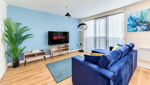 Un salon avec un canapé bleu, un téléviseur affichant le logo « NETFLIX », une lampe sur pied et une grande plante.