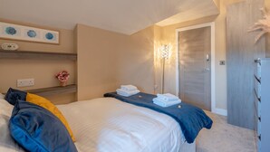 A hotel room with a bed, a bedside table with a vase of flowers, a wall-mounted clock, and a door with a decorative panel. - Moat House Penthouse Suite - 2 Bed  2 Bath (Rawtenstall)