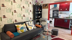 Un salon avec un canapé gris, des coussins colorés, une table basse en verre et un tapis rouge. À côté se trouve une cuisine avec des placards rouges, un lave-linge et une carte du monde accrochée au mur.