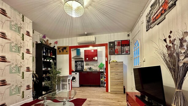 Un salon moderne avec une table basse en verre, un meuble de rangement rouge et un téléviseur. On aperçoit une cuisine à l’arrière-plan, avec des placards rouges et une carte du monde accrochée au mur.