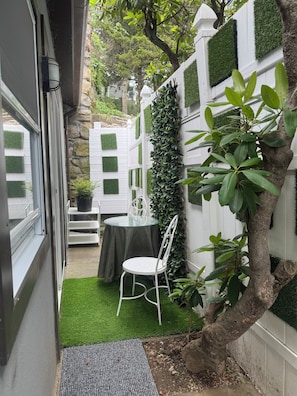 Uma pequena área externa com mesas e cadeiras, cercada por um muro branco com áreas verdes, uma árvore e uma parede branca.