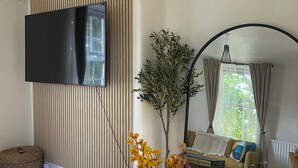 A living room with a flat-screen TV mounted on the wall, a sofa with cushions, a small round table, and a potted plant.