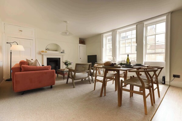 A spacious living room with a fireplace, a sofa, a dining table with chairs, and large windows.
