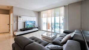 Una sala de estar moderna con un televisor de pantalla plana grande, sofás grises, una mesa de centro de vidrio y un balcón con mobiliario exterior.