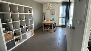 Ein geräumiger Raum mit einer großen weißen Regalkonstruktion, gefüllt mit verschiedenen Gegenständen, einer Essecke mit Holztisch und -stühlen sowie Blick durch ein Fenster mit transparenten Vorhängen.