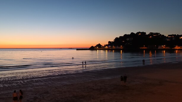 Una playa al atardecer con aguas tranquilas, algunas personas caminando y edificios al fondo.