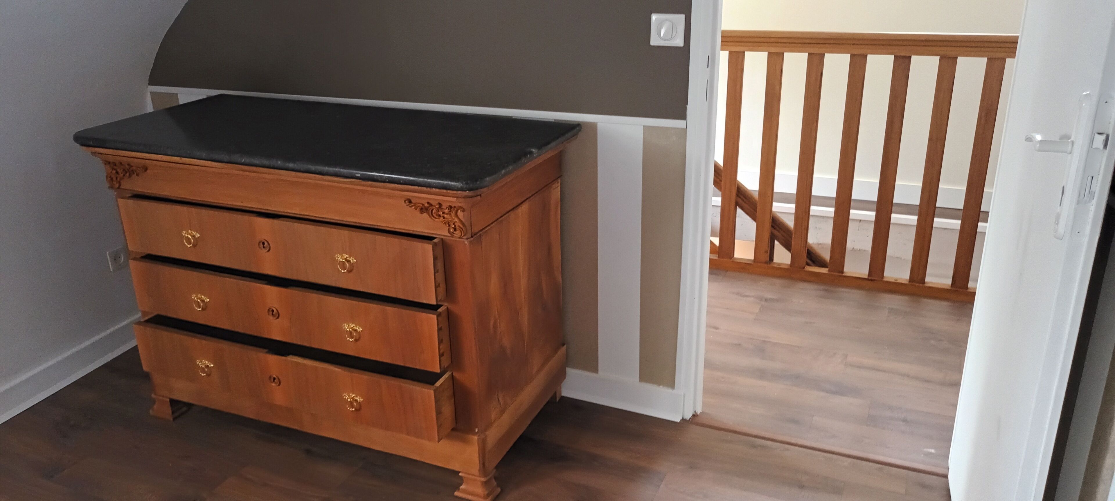 Une commode en bois, avec un plateau noir et trois tiroirs, située dans une pièce dont le sol est recouvert de parquet et dont un des murs est blanc.
