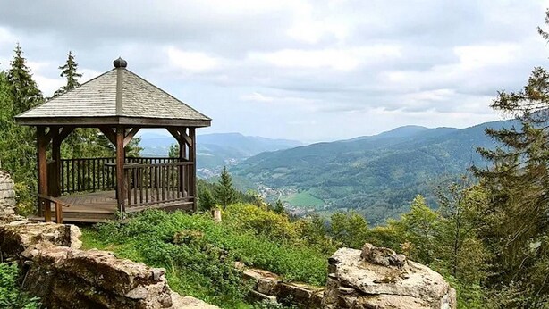 Un belvédère en bois, doté d’une balustrade en métal, est entouré d’une végétation luxuriante et d’un terrain rocheux, et offre une vue sur une vallée et des montagnes lointaines.