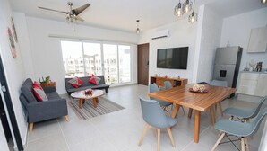 A modern living room with a sofa, coffee table, and dining table with chairs. There is a TV mounted on the wall and a ceiling fan. - Close to the Turquoise Sea, ideal for couples, adventurers and families! (Cancún)