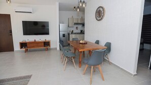 A modern living room with a wooden dining table, grey chairs, a flat-screen TV, and a kitchen in the background. - Close to the Turquoise Sea, ideal for couples, adventurers and families! (Cancún)