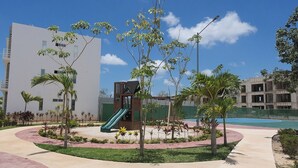 A playground with a slide, surrounded by palm trees and a basketball court, with buildings in the background. - Close to the Turquoise Sea, ideal for couples, adventurers and families! (Cancún)