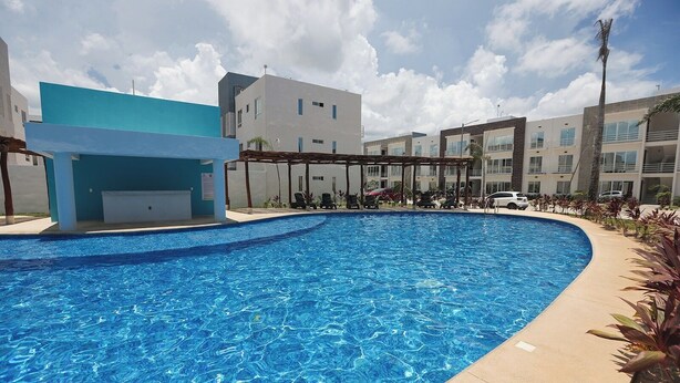 A swimming pool with a curved edge, surrounded by a covered area with chairs and a building in the background. - Close to the Turquoise Sea, ideal for couples, adventurers and families! (Cancún)